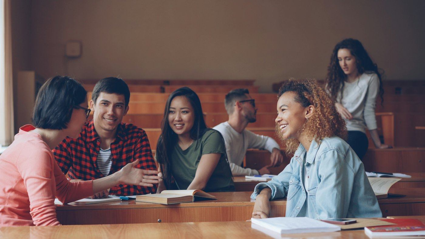 Grupo de estudiantes conversando en un aula universitaria con libros y apuntes sobre la mesa