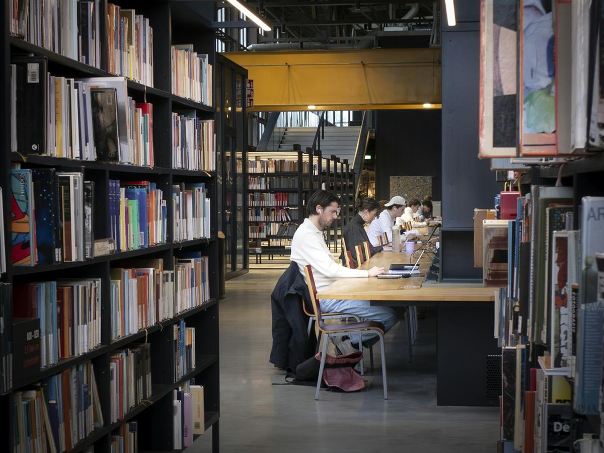 Estudiantes trabajando con sus portátiles en una mesa larga de una biblioteca, entre estanterías de libros